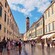 People walking on a pedestrian street in a medieval town lined with shops and a bell tower
